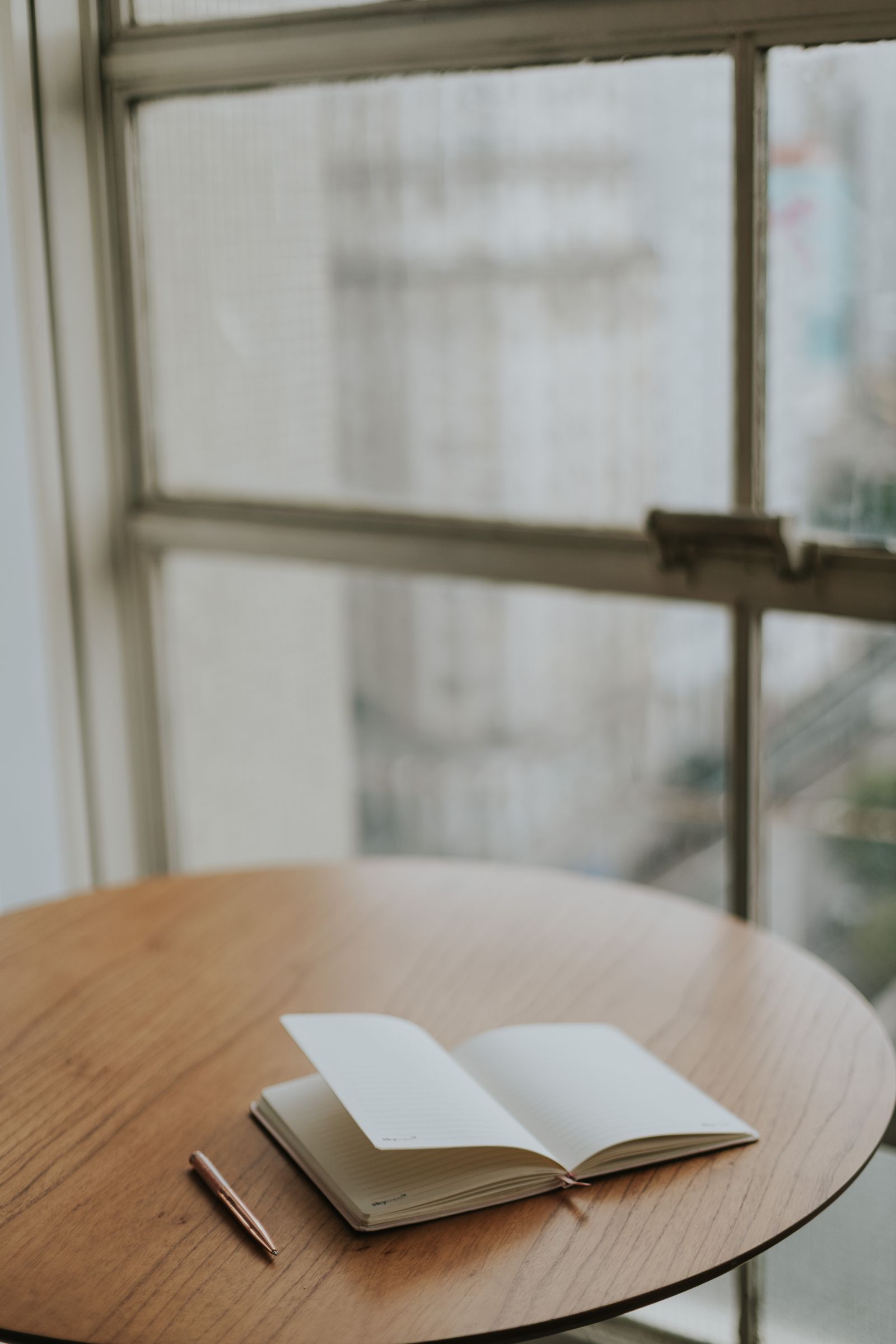 An open journal with a pen on a wooden table by a window, offering a cozy writing space.